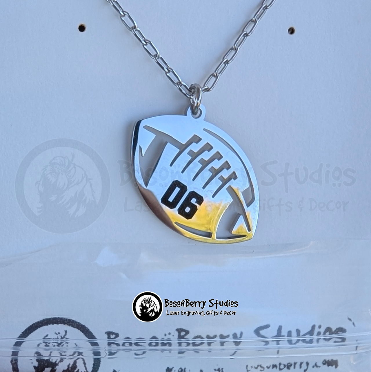 Silver football-shaped pendant on a chain with '06' engraved on it in black varsity-style font, on a white jewelry display card, featuring 'BosonBerry Studios' logo.