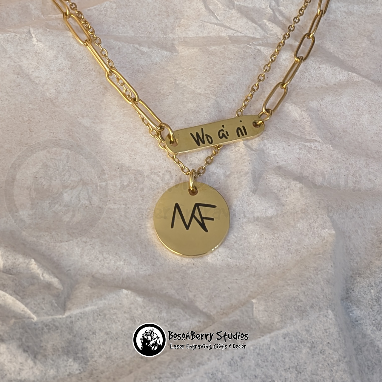 Two gold necklaces, one a paperclip chain with an oval pendant engraved with 'Wo ai ni', and one a small round pendant engraved with a stylized monogram 'NAF', on a crinkled white paper background with 'BosonBerry Studios' branding.