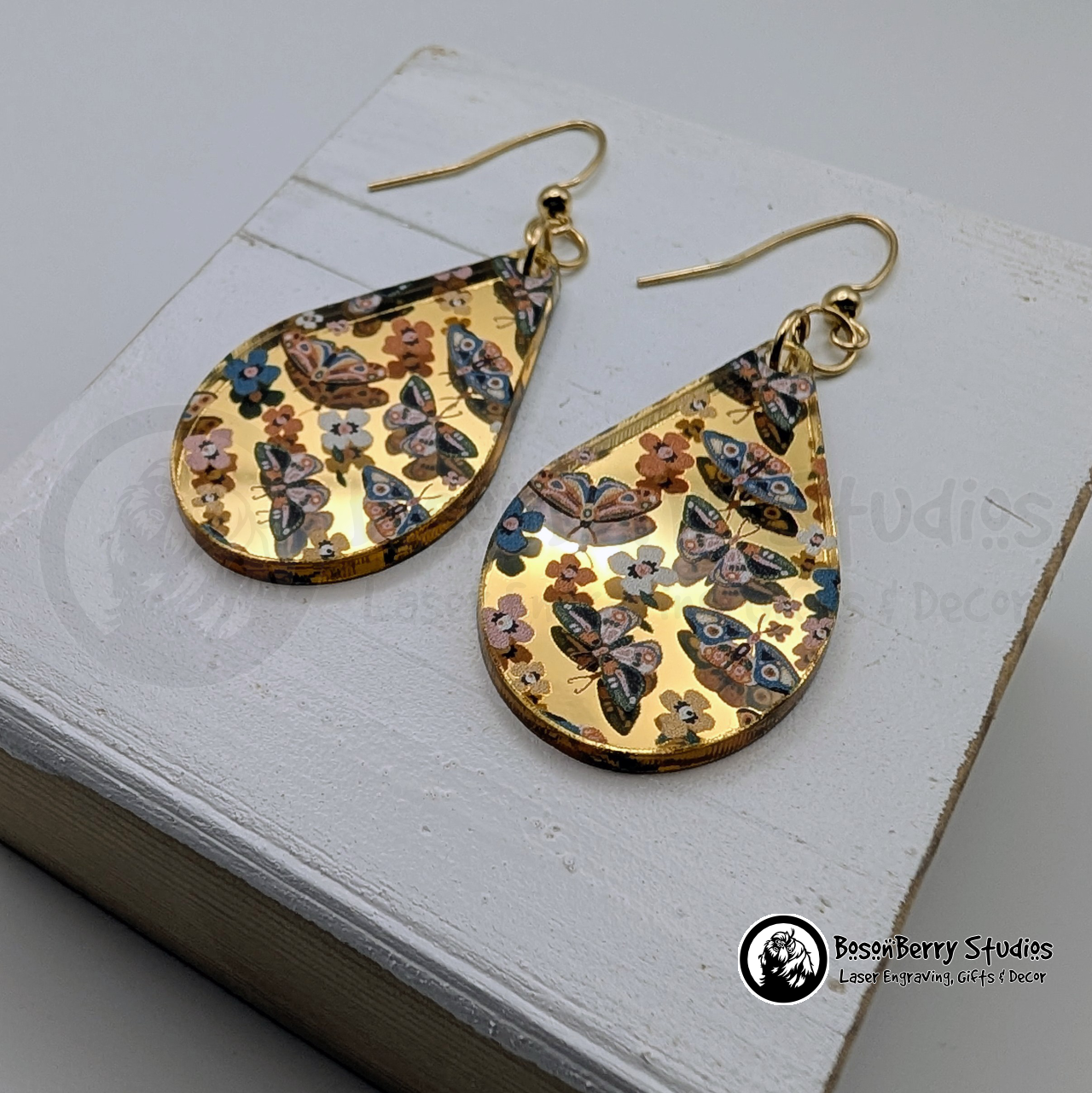 Teardrop shaped gold-mirrored acrylic earrings with a colorful butterflies & moths pattern, on a wooden stand with 'BosonBerry Studios' branding.