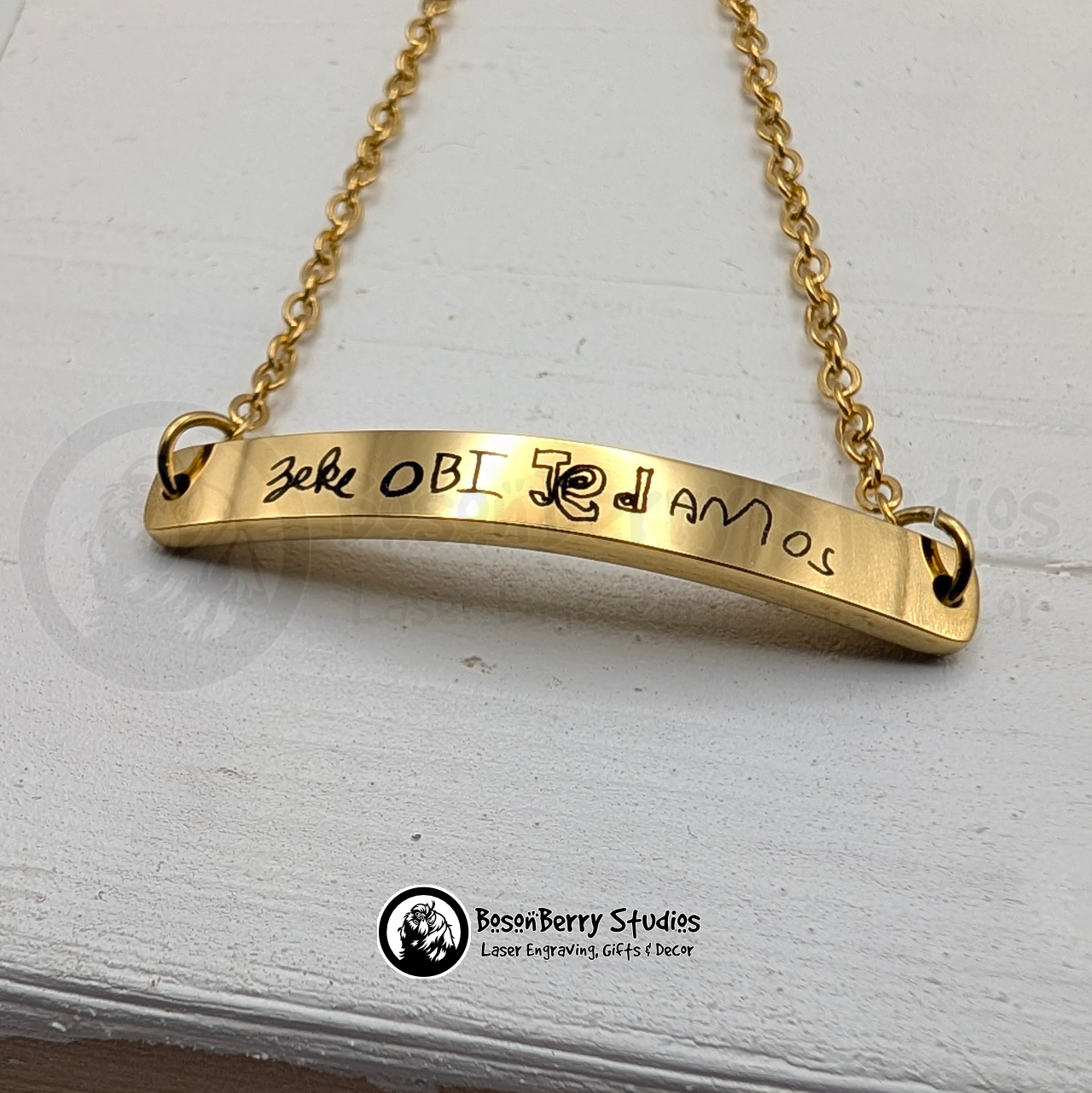Gold bar bracelet, engraved with black handwritten names, on a white wooden stand with 'BosonBerry Studios' branding.