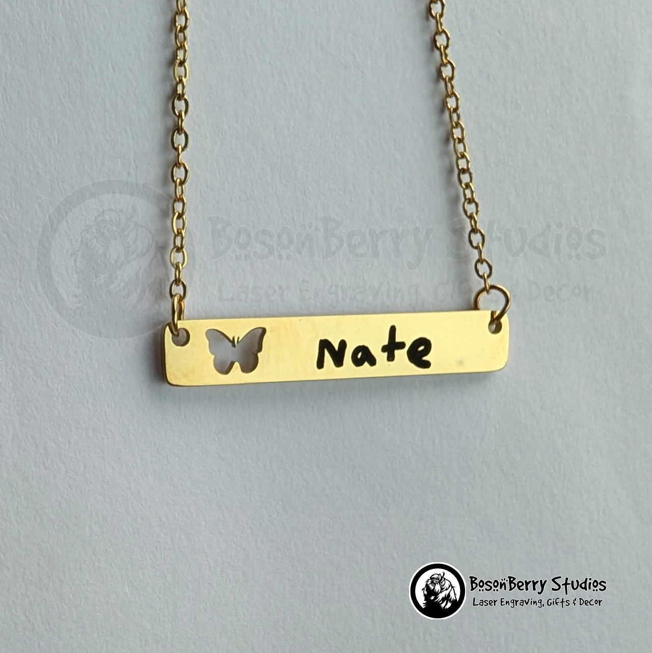 Gold bar necklace with a butterfly cutout and 'Nate' engraved in black handwriting on a white jewelry display card with 'BosonBerry Studios' branding.