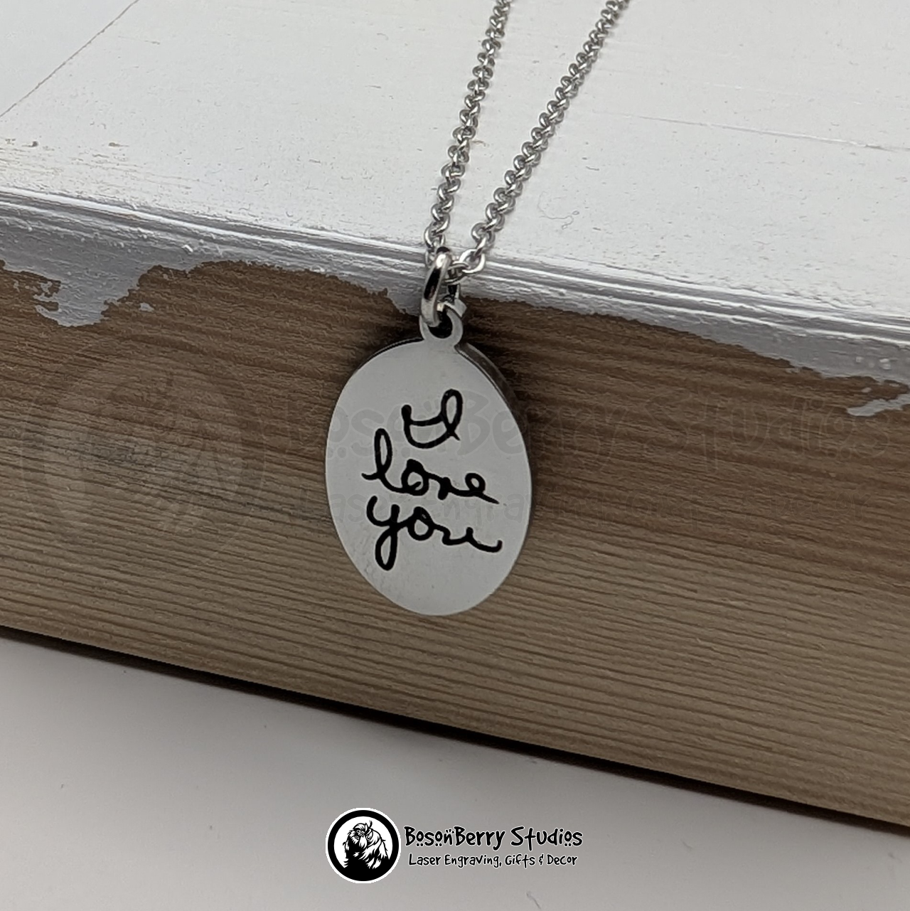 Silver oval pendant necklace, engraved with black handwriting that says "I love you" in cursive, hanging on a white wooden stand with 'BosonBerry Studios' branding.