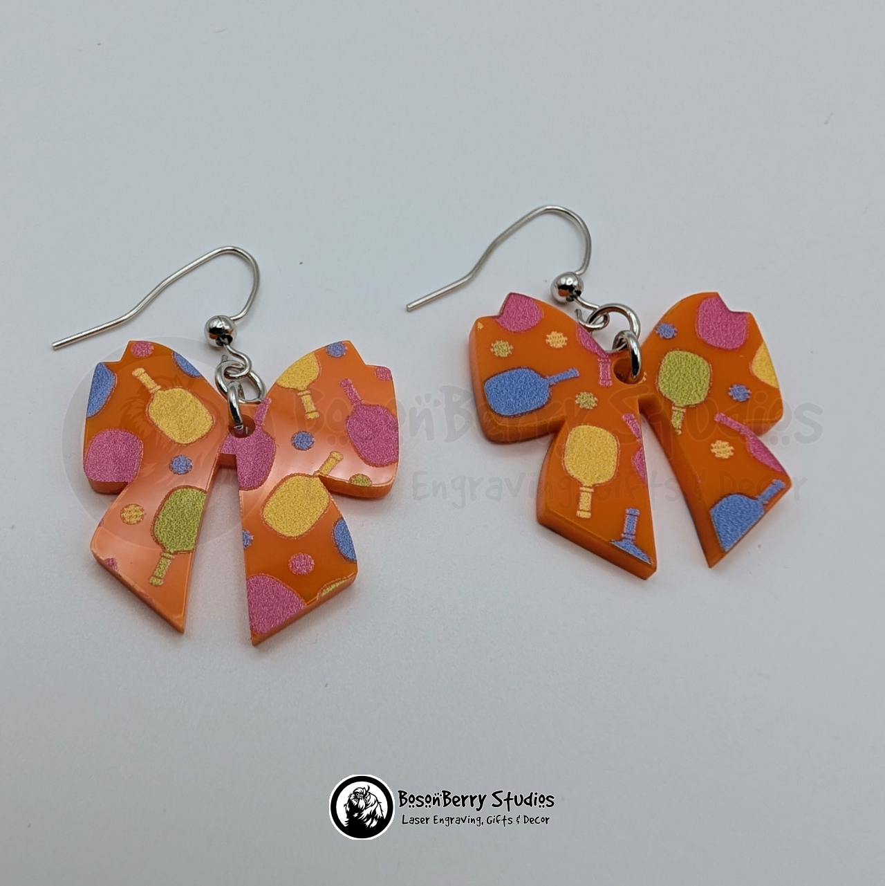 Orange bow-shaped acrylic earrings with a colorful pickleball pattern on a white background with 'BosonBerry Studios' branding.