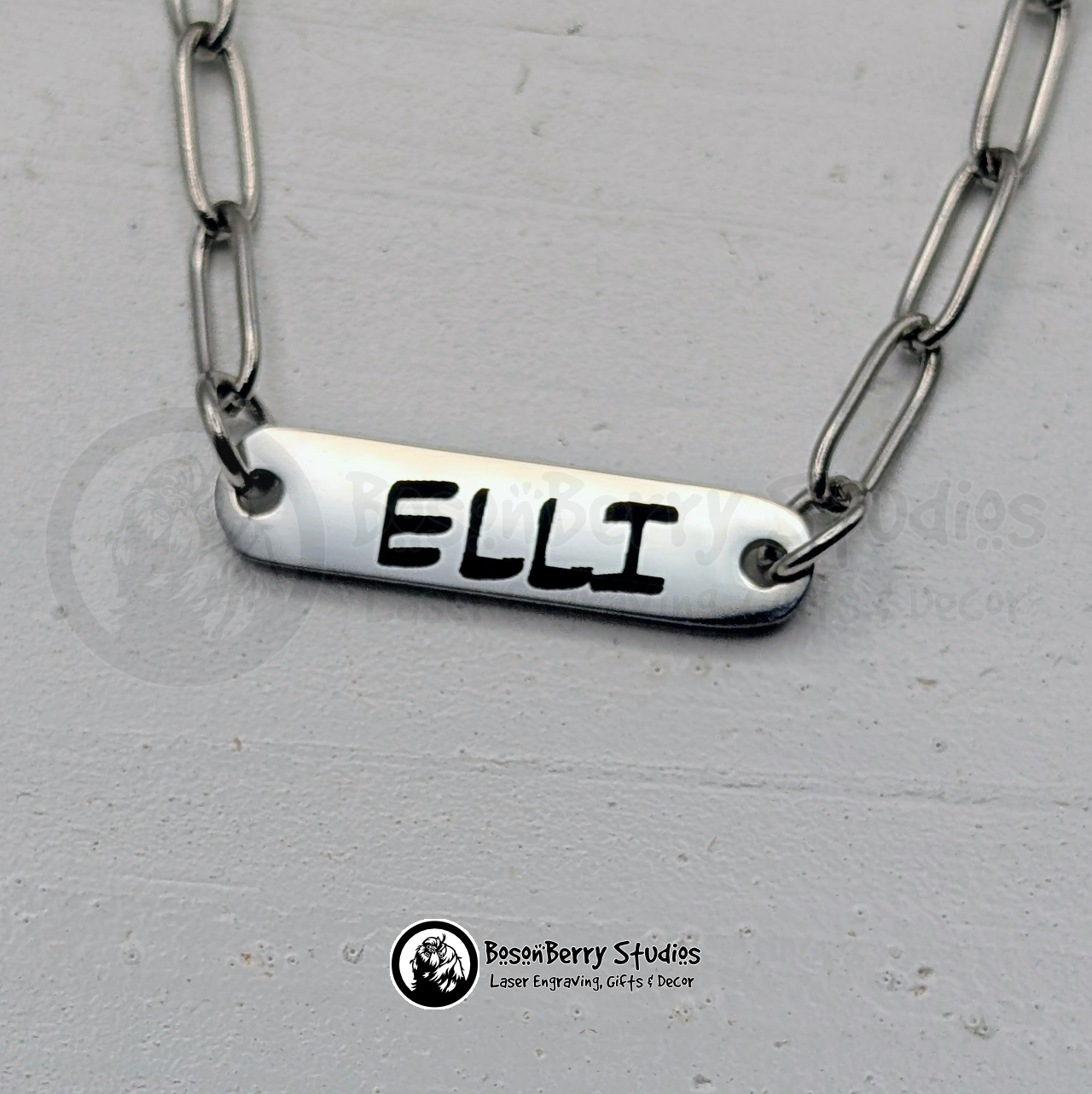 Silver paperclip chain necklace with oval bar pendant engraved with 'ELLI' in a black, printed font, on a white wooden stand with 'BosonBerry Studios' branding.