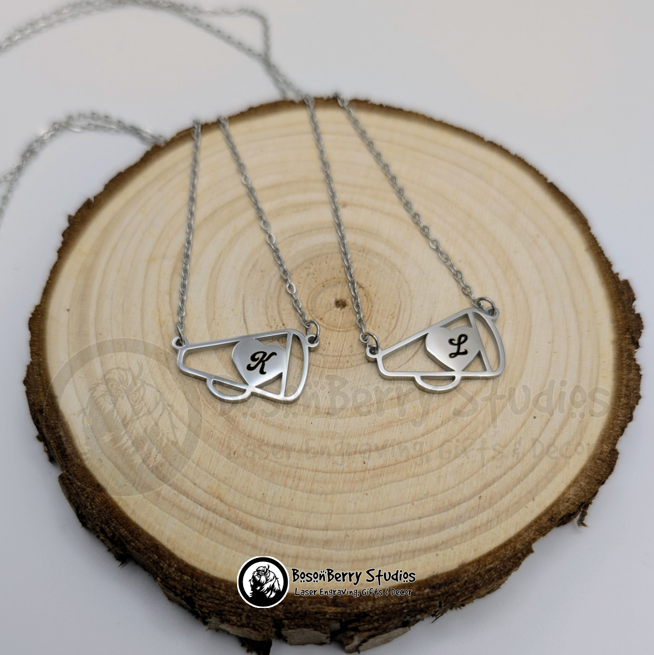Two silver cheerleader megaphone necklaces with cursive letter 'K' and 'L' engraved on them, on a wooden surface, featuring 'BosonBerry Studios' branding.