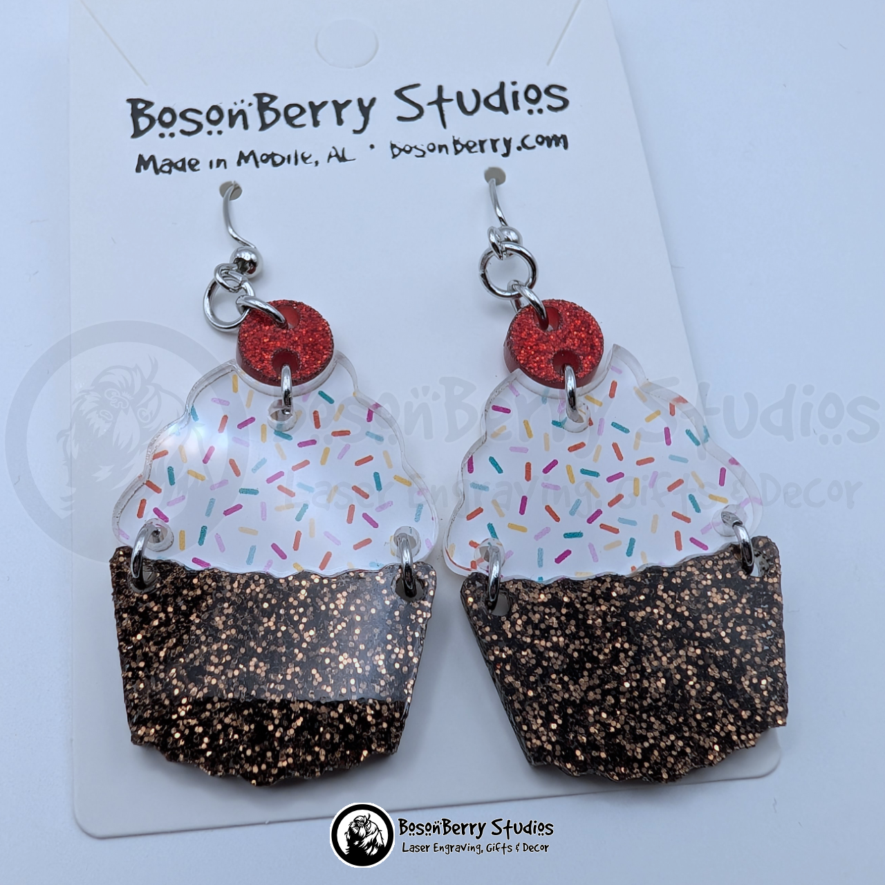 Tiered acrylic earrings with a glitter chocolate base, a white with colored sprinkles icing center, and a red glitter cherry top, on a BosonBerry Studios branded display card.