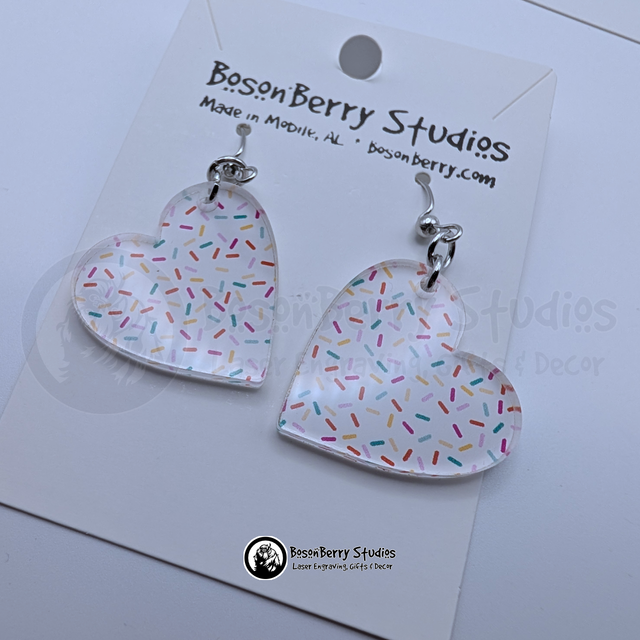 White heart-shaped earrings with a colorful sprinkles pattern, on cards with BosonBerry Studios branding.