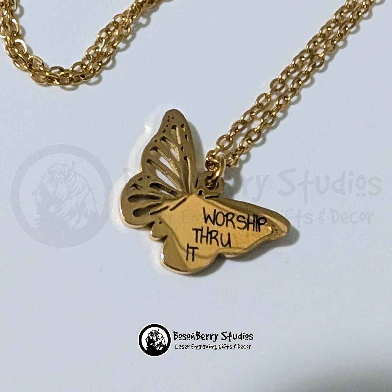 Gold butterfly necklace with words 'Worship Thru It' engraved in black, printed font on a white background with 'BosonBerry Studios' branding.