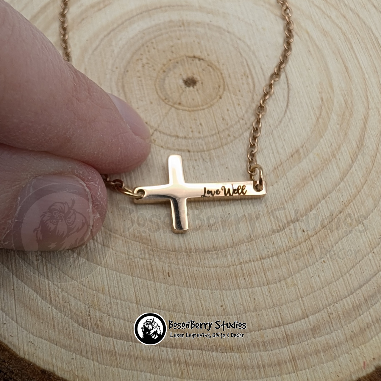 Rose gold necklace with a tiny cross pendant engraved with 'Love Well' in black cursive letters, displayed on a wooden surface next to a fingertip for scale, featuring 'BosonBerry Studios' branding.