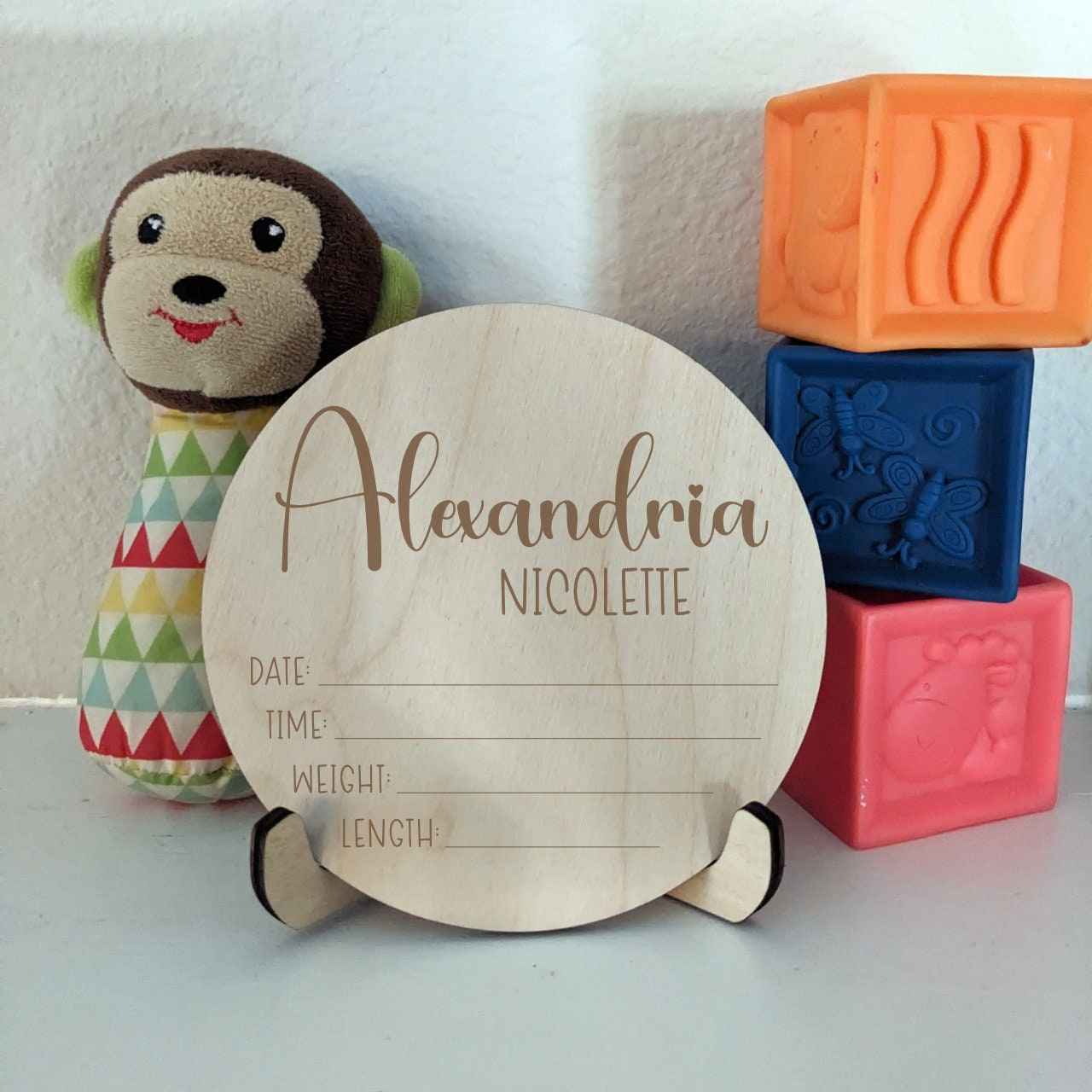 Baby Name Birth Stats 5" Round Sign displayed in easel with toy monkey and bright blocks in background.