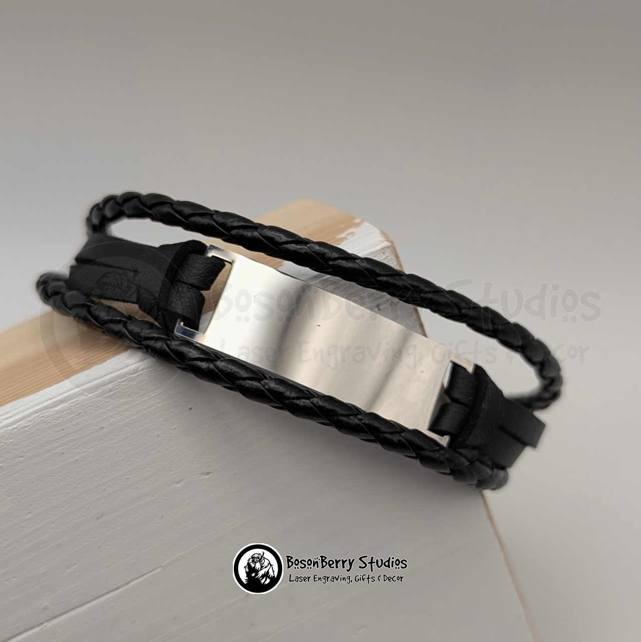 Black braided triple cord bracelet with a silver bar pendant on a white wooden stand with 'BosonBerry Studios' branding.