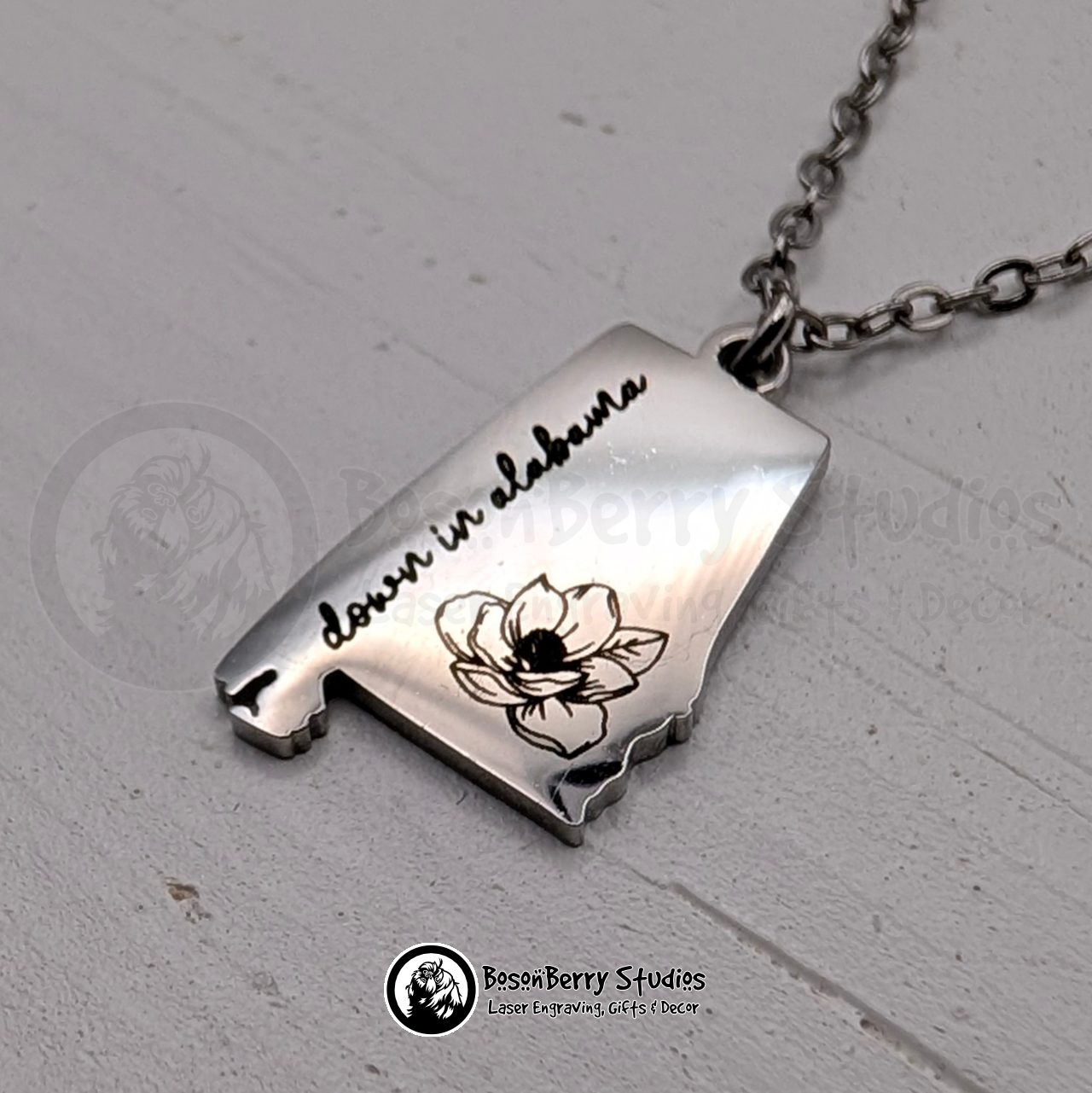Silver necklace with an Alabama state-shaped pendant, engraved with a magnolia flower design and 'down in Alabama' text in cursive, on a white wooden stand with 'BosonBerry Studios' branding.