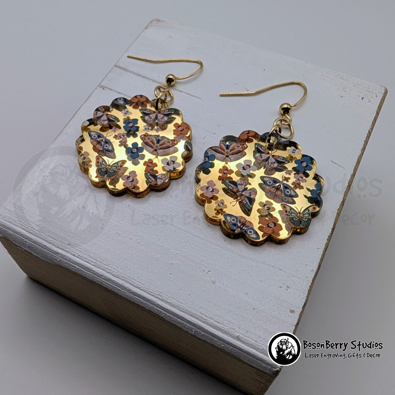 Scalloped Circle shaped gold-mirrored acrylic earrings with a colorful butterflies & moths pattern, on a wooden stand with 'BosonBerry Studios' branding. 