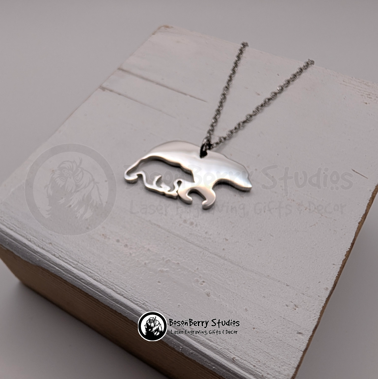 A silver bear-shaped pendant necklace with cub-shaped cutout on a white wooden stand with 'BosonBerry Studios' branding.