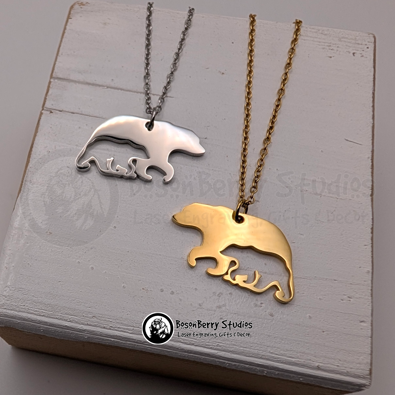 Two bear-shaped pendant necklaces with cub shaped cutouts, one silver and one gold, on a white wooden stand with 'BosonBerry Studios' branding.