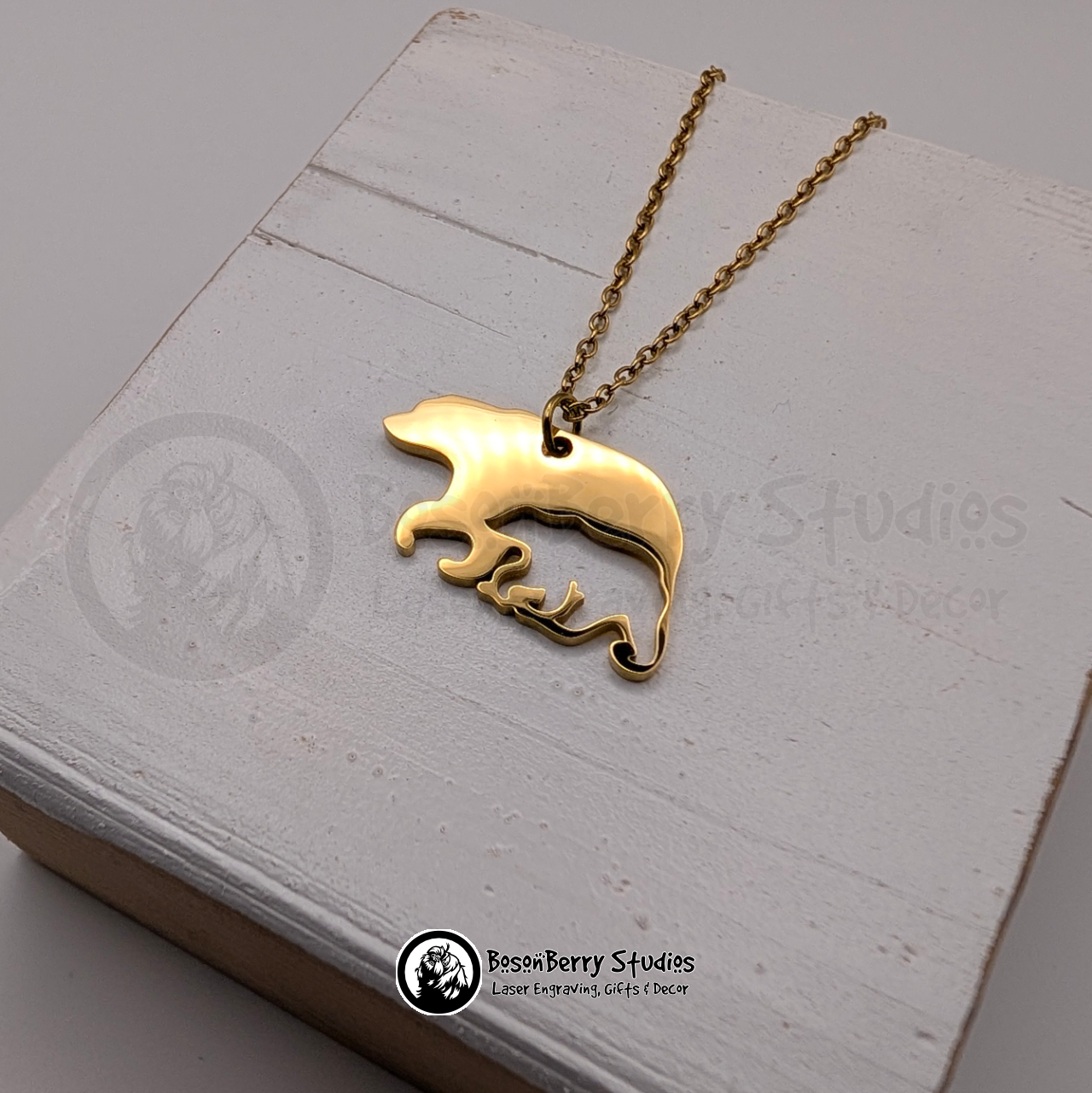 A gold bear-shaped pendant necklace with cub-shaped cutout on a white wooden stand with 'BosonBerry Studios' branding.