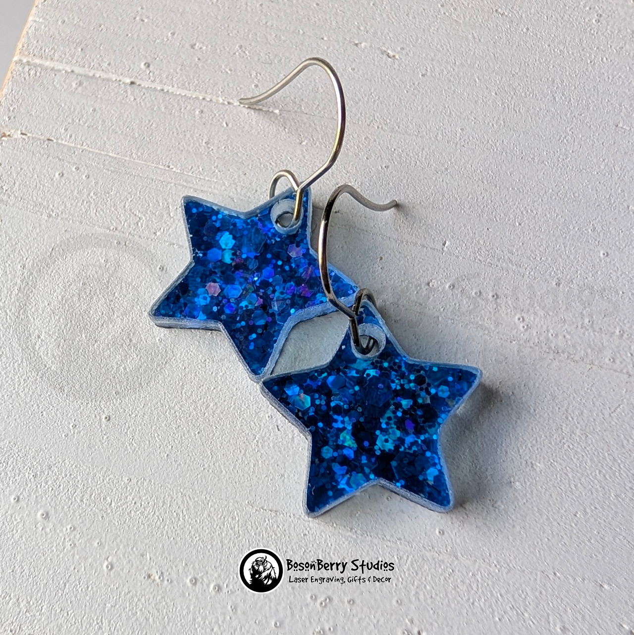 Star-shaped blue glitter earrings on a white background, branded 'BosonBerry Studios'.