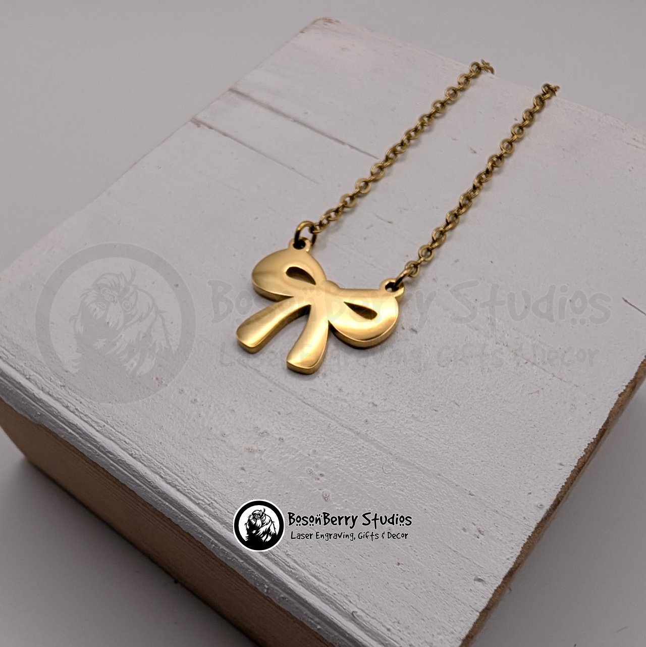 Gold bow-shaped pendant necklace on a white wooden stand with 'BosonBerry Studios' branding.