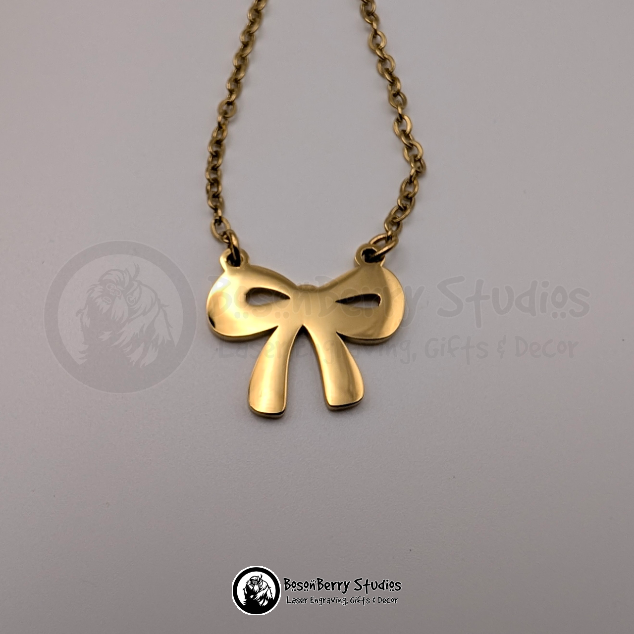 Gold bow-shaped pendant necklace on a white background with 'BosonBerry Studios' branding.