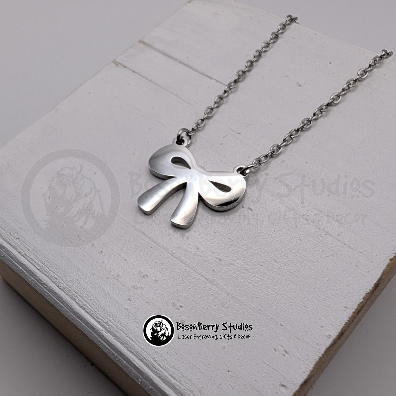 Silver necklace with a bow pendant on a white wooden stand with 'BosonBerry Studios' branding.