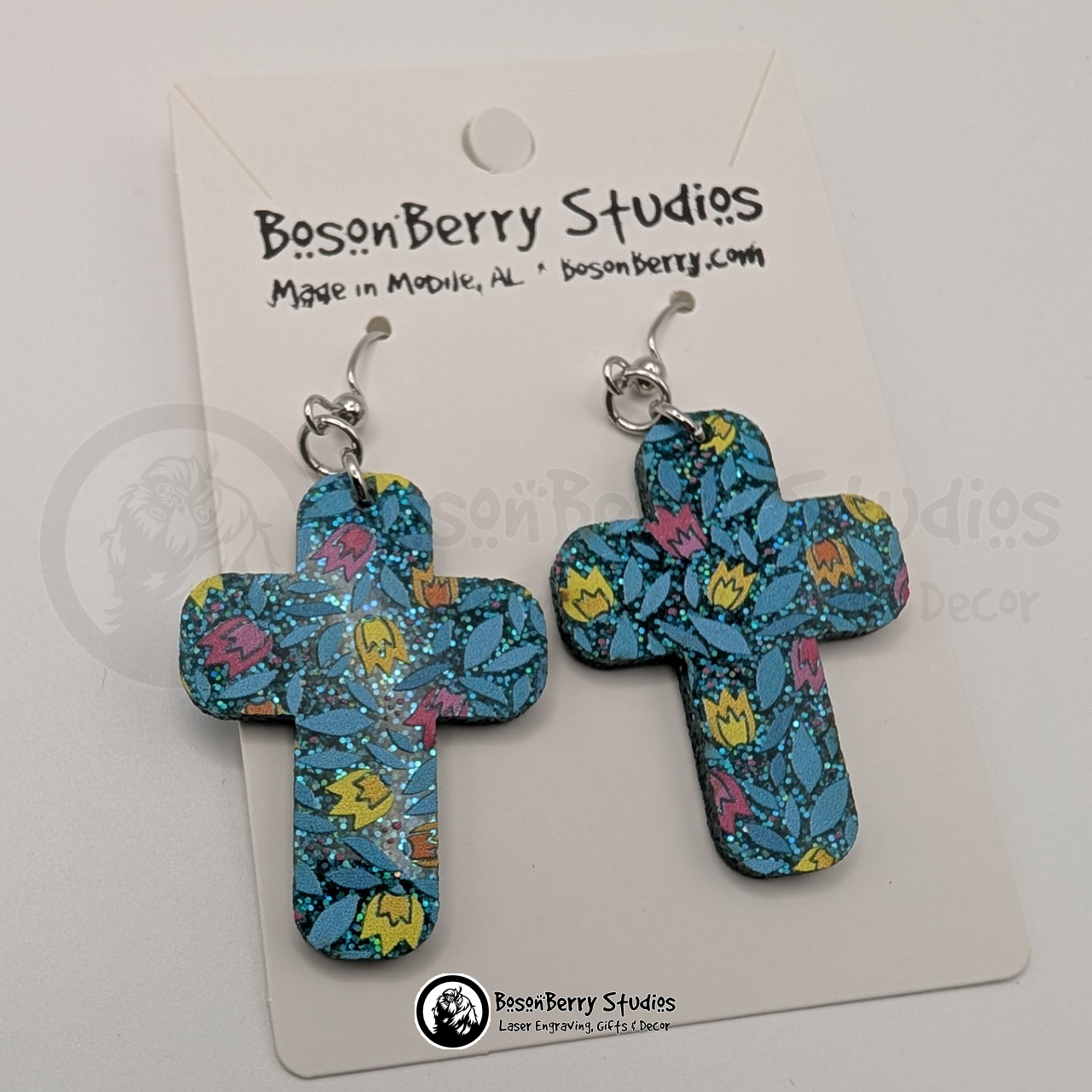 Earrings featuring a pattern of cartoon tulips in pink, yellow and orange on a teal blue glitter field, in a large cross shape, on display cards with 'BosonBerry Studios' branding.