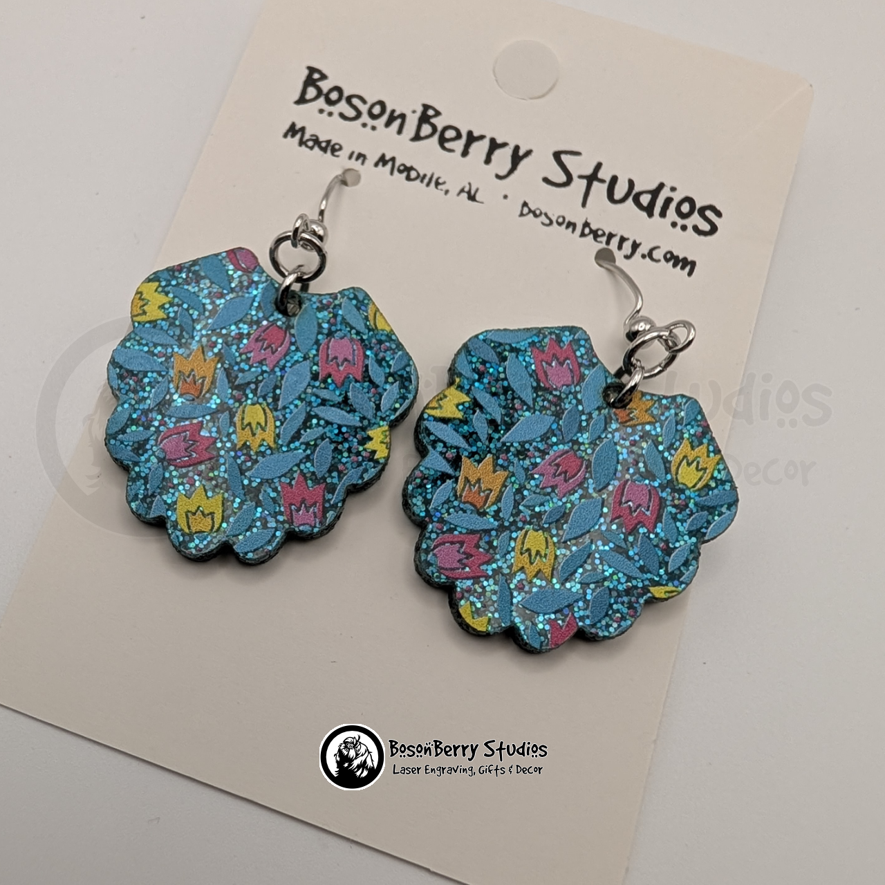 Earrings featuring a pattern of cartoon tulips in pink, yellow and orange on a teal blue glitter field, in a scalloped fan shape, on display cards with 'BosonBerry Studios' branding.