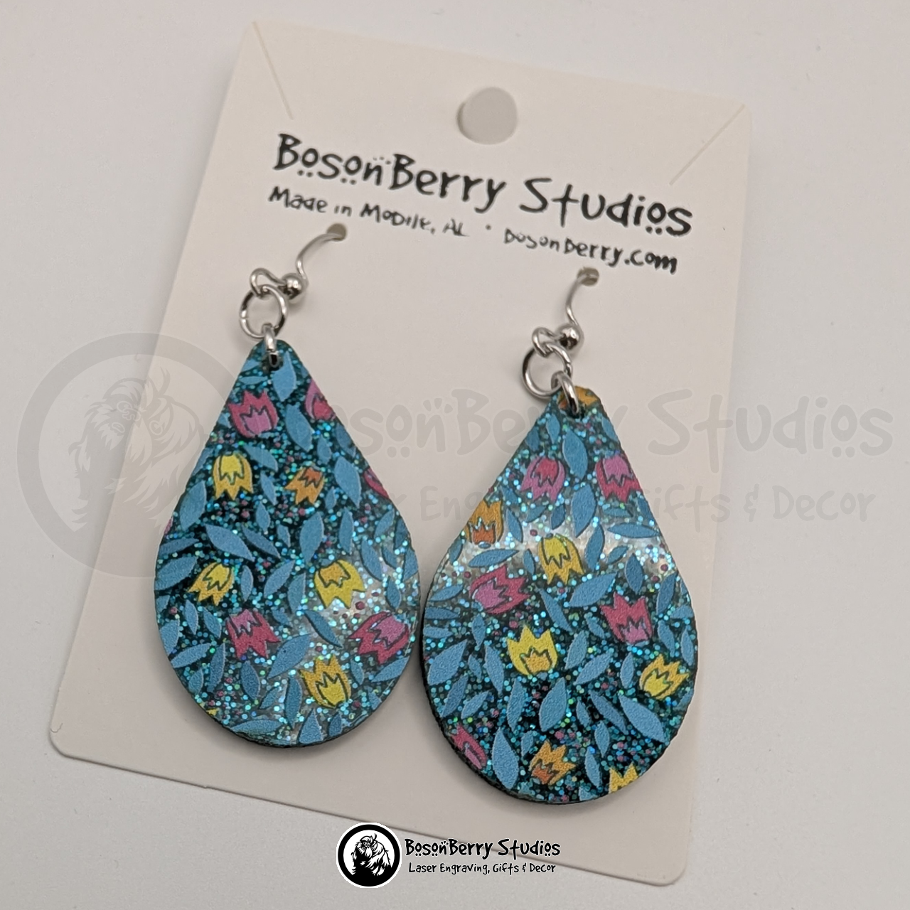 Earrings featuring a pattern of cartoon tulips in pink, yellow and orange on a teal blue glitter field, in a teardrop shape, on display cards with 'BosonBerry Studios' branding.