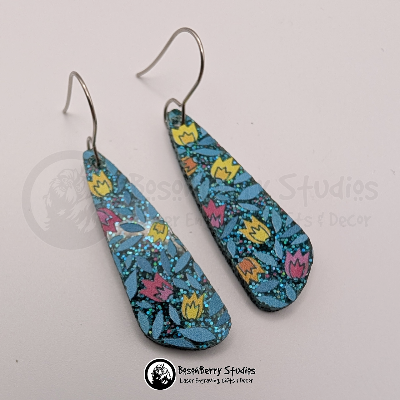 Earrings featuring a pattern of cartoon tulips in pink, yellow and orange on a teal blue glitter field, in a skinny, elongated teardrop shape, on a white background with 'BosonBerry Studios' branding.