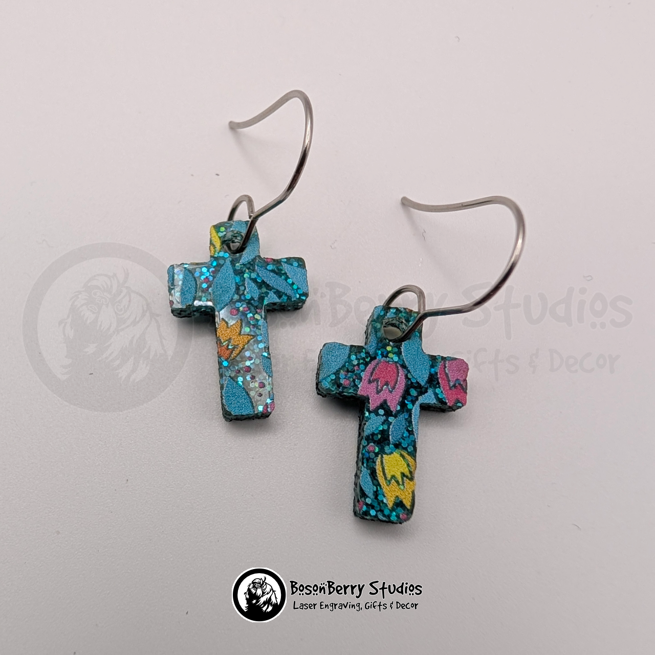 Earrings featuring a pattern of cartoon tulips in pink, yellow and orange on a teal blue glitter field, in a tiny cross shape, on a white background with 'BosonBerry Studios' branding.