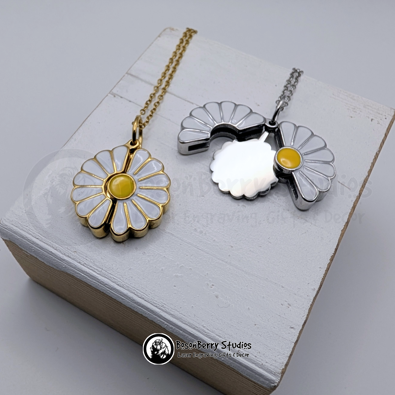 Two white enameled Daisy Reveal pendants, one gold and one silver, on a white wooden stand with 'BosonBerry Studios' branding.