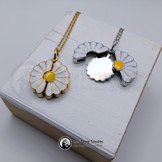 Two white enameled Daisy Reveal pendants, one gold and one silver, on a white wooden stand with 'BosonBerry Studios' branding.