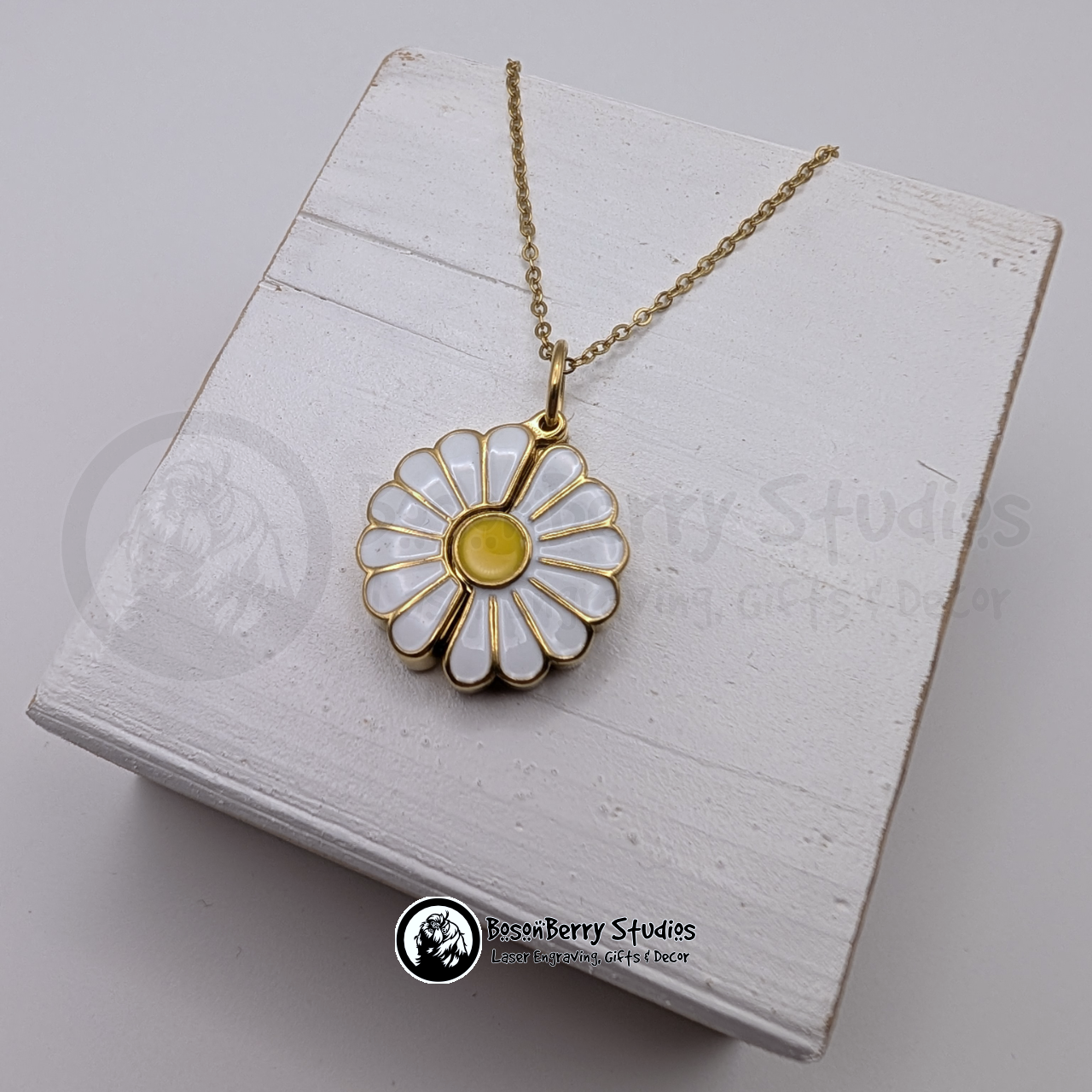 Gold necklace with a white and yellow enameled daisy pendant on a white wooden stand with 'BosonBerry Studios' branding.