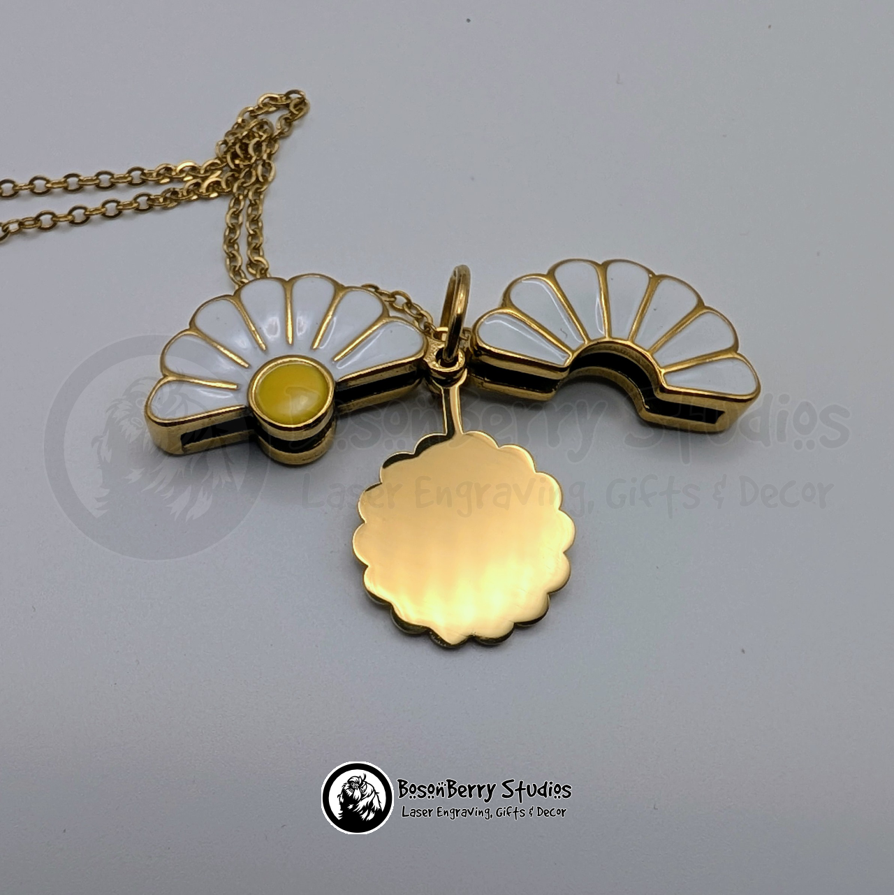 Gold necklace with a white enameled daisy pendant, opened to reveal a scalloped gold disc, on a white background with 'BosonBerry Studios' branding.