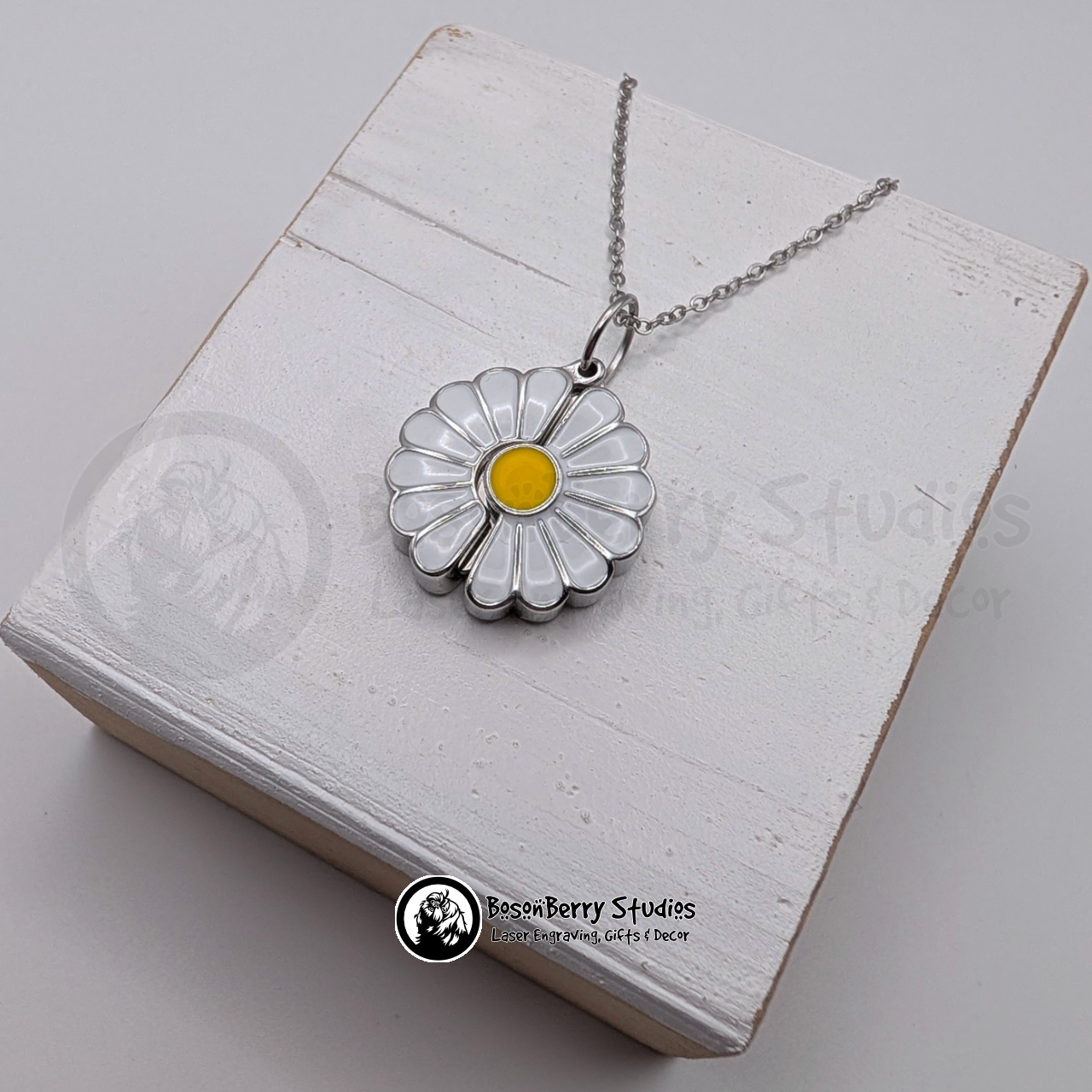 Silver necklace with white enameled daisy-shaped pendant on a white wooden stand with 'BosonBerry Studios' branding.