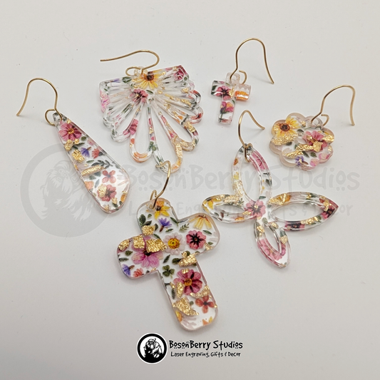 Clear acrylic earrings with embedded gold flakes and a multicolor floral print, in multiple shapes included cross, half daisy, butterfly and flower, on a white background with 'BosonBerry Studios' branding.