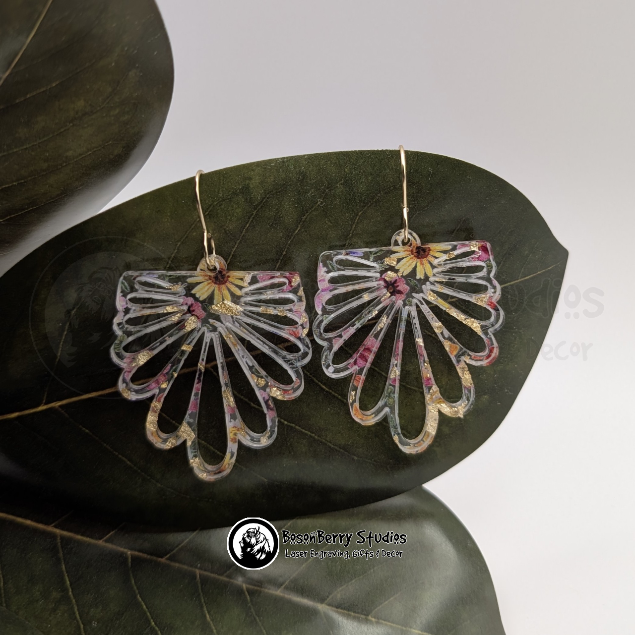 Clear acrylic earrings with embedded gold flakes and a multicolor floral print, in a floral petal burst half daisy shape, on a green magnolia leaf in front of a white background with 'BosonBerry Studios' branding.