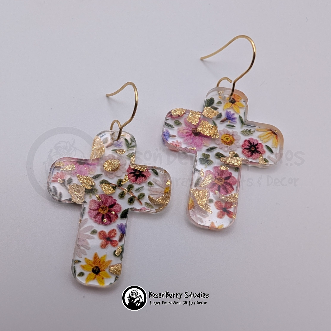 Clear acrylic earrings with embedded gold flakes and a multicolor floral print, in a large cross shape, on a white background with 'BosonBerry Studios' branding.