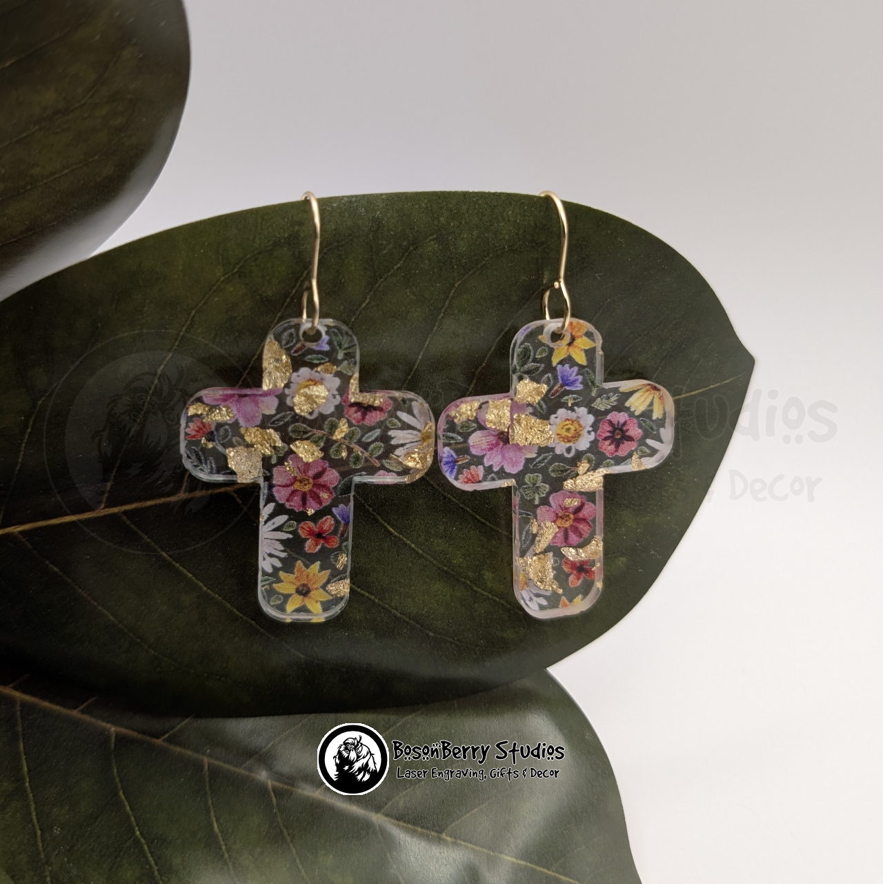 Clear acrylic earrings with embedded gold flakes and a multicolor floral print, in a large cross shape, on a green magnolia leaf in front of a white background with 'BosonBerry Studios' branding.