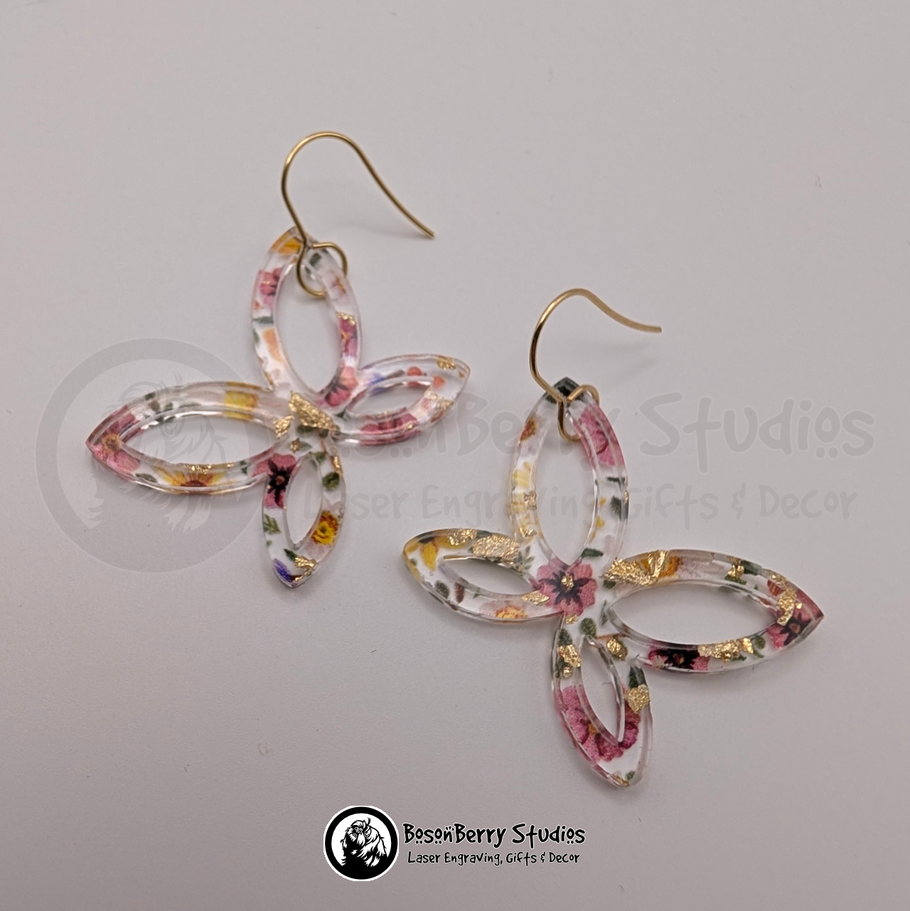 Clear acrylic earrings with embedded gold flakes and a multicolor floral print, in an abstract butterfly shape, on a white background with 'BosonBerry Studios' branding.
