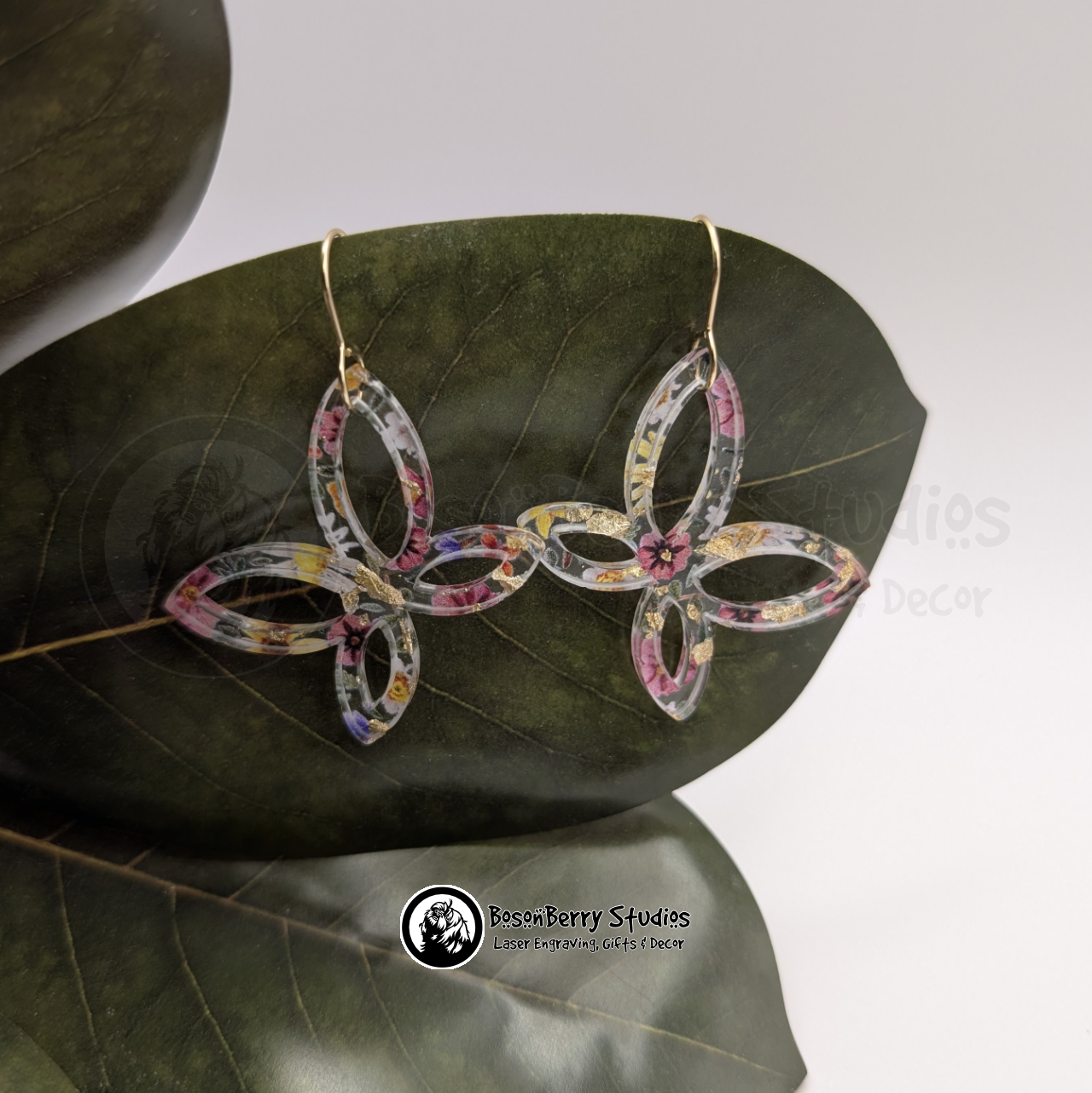 Clear acrylic earrings with embedded gold flakes and a multicolor floral print, in an abstract butterfly shape, on a green magnolia leaf in front of a white background with 'BosonBerry Studios' branding.