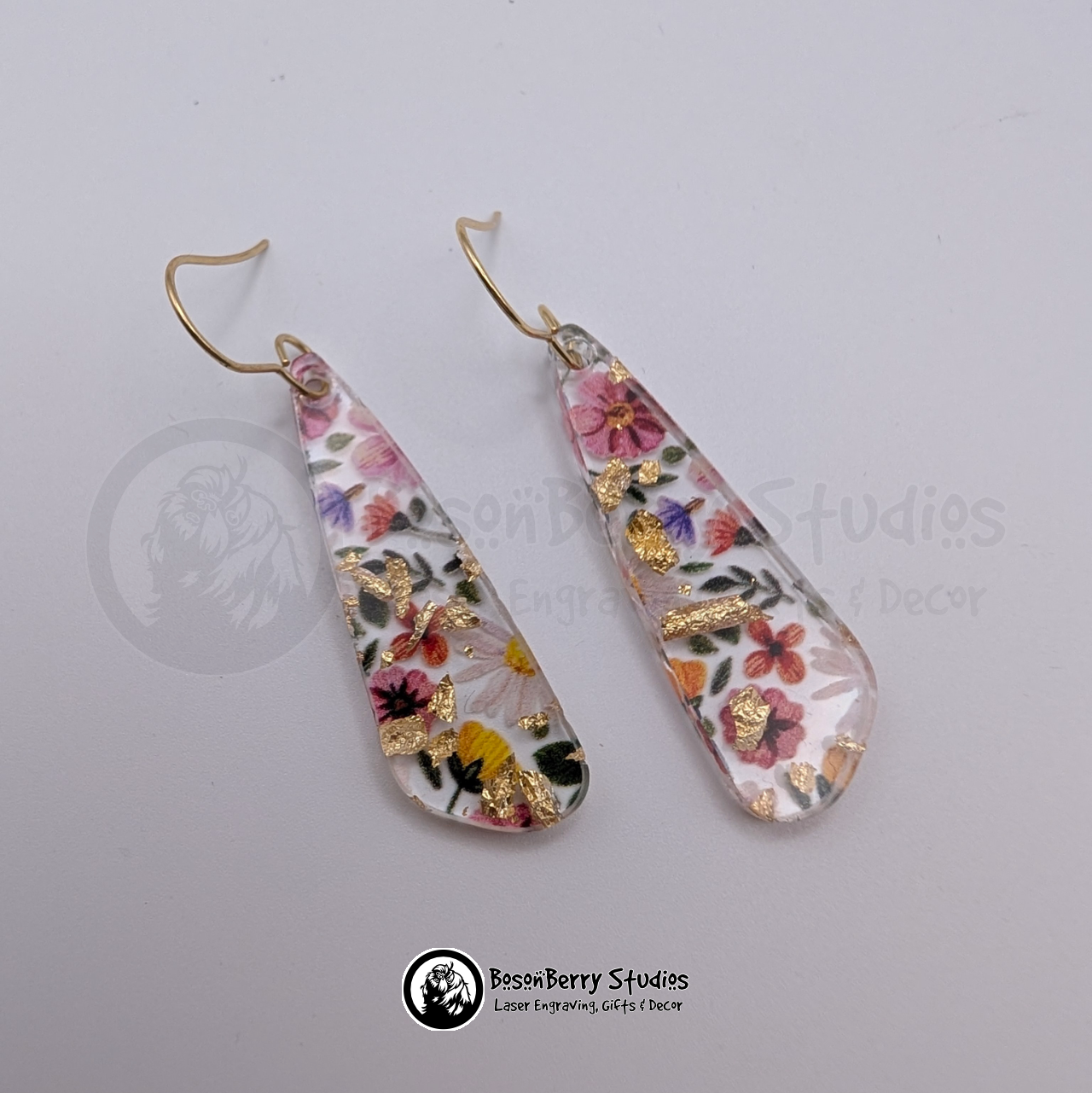 Clear acrylic earrings with embedded gold flakes and a multicolor floral print, in a skinny teardrop shape, on a white background with 'BosonBerry Studios' branding.