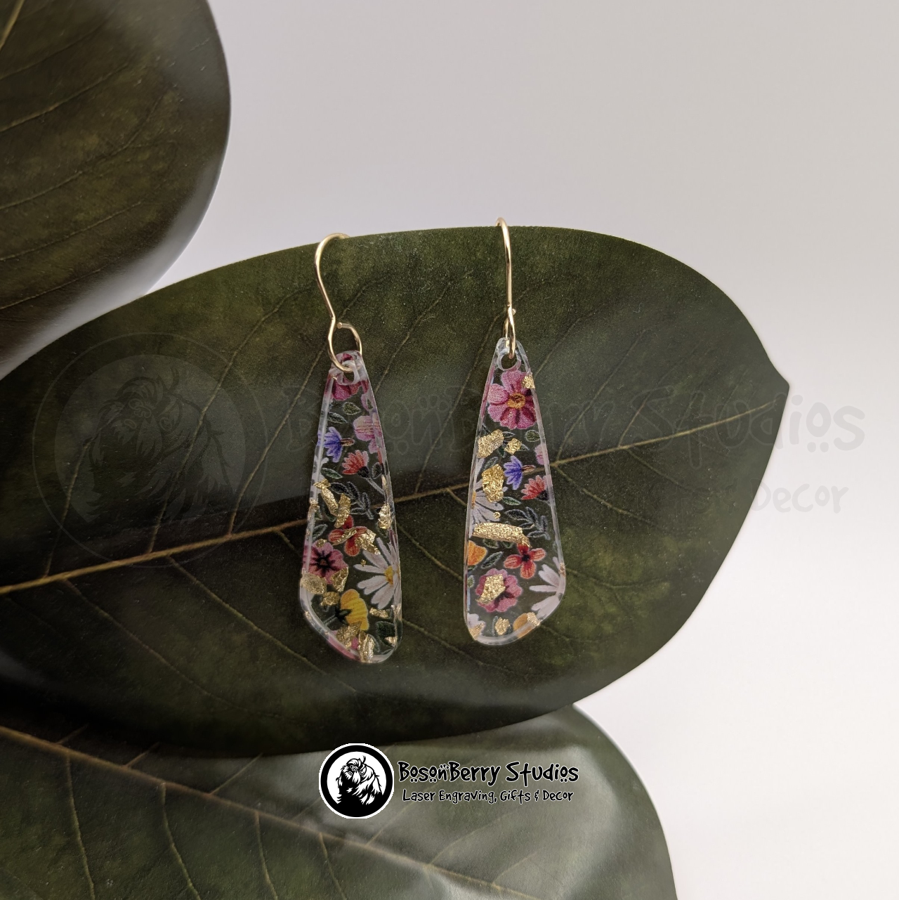 Clear acrylic earrings with embedded gold flakes and a multicolor floral print, in a skinny teardrop shape, on a green magnolia leaf in front of a white background with 'BosonBerry Studios' branding.
