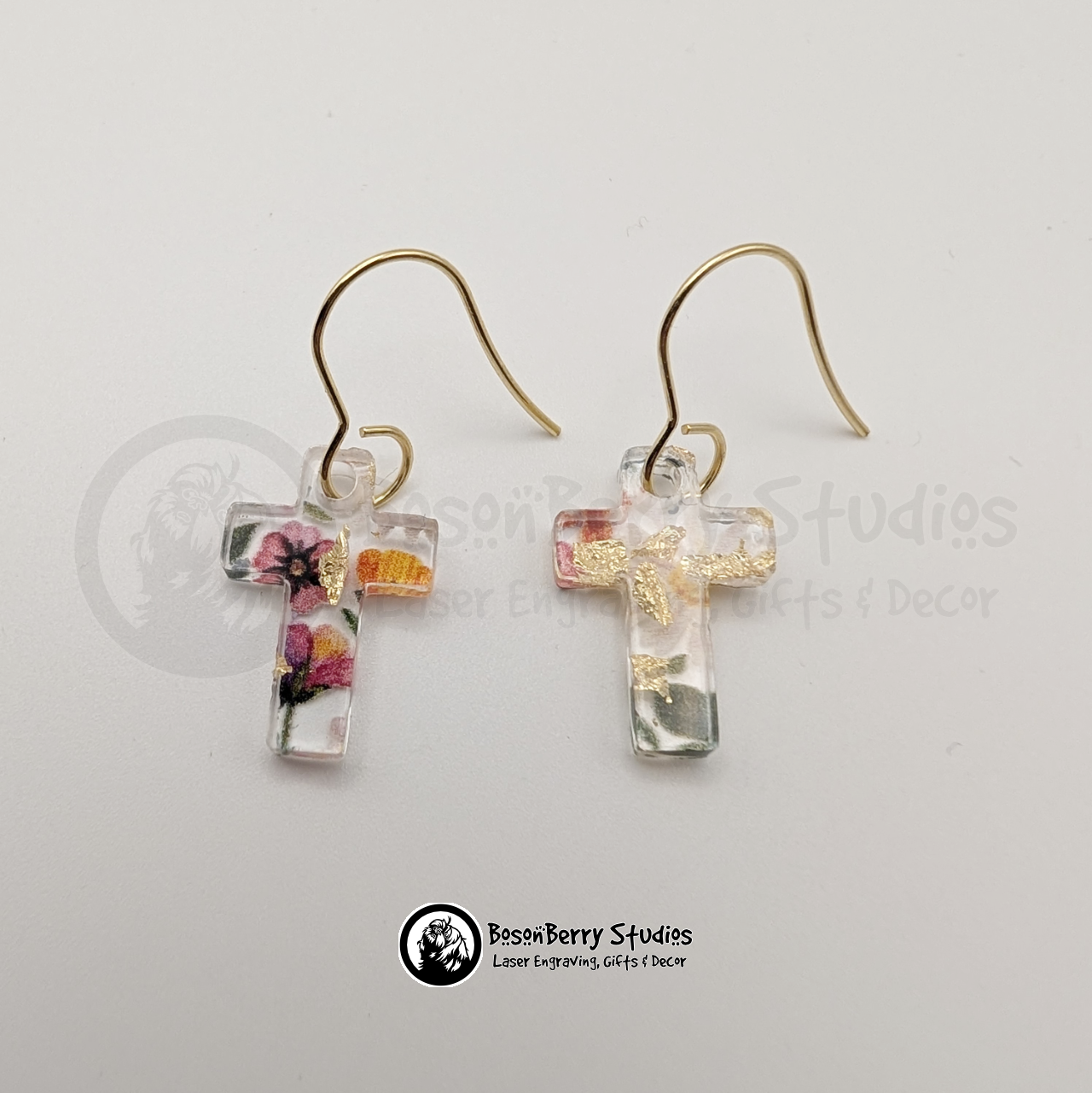 Clear acrylic earrings with embedded gold flakes and a multicolor floral print, in a tiny cross shape, on a white background with 'BosonBerry Studios' branding.