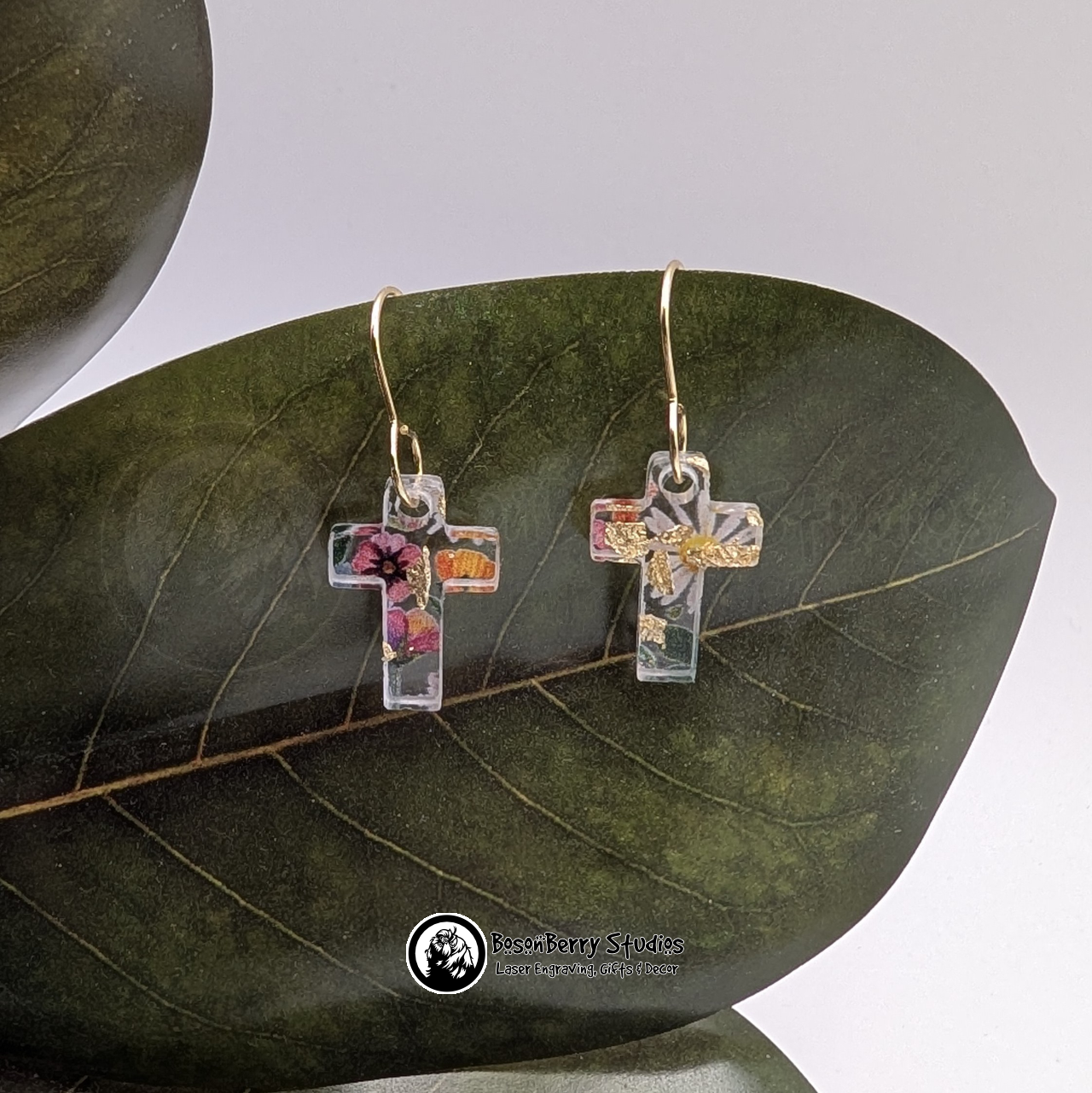 Clear acrylic earrings with embedded gold flakes and a multicolor floral print, in a tiny cross shape, on a green magnolia leaf in front of a white background with 'BosonBerry Studios' branding.
