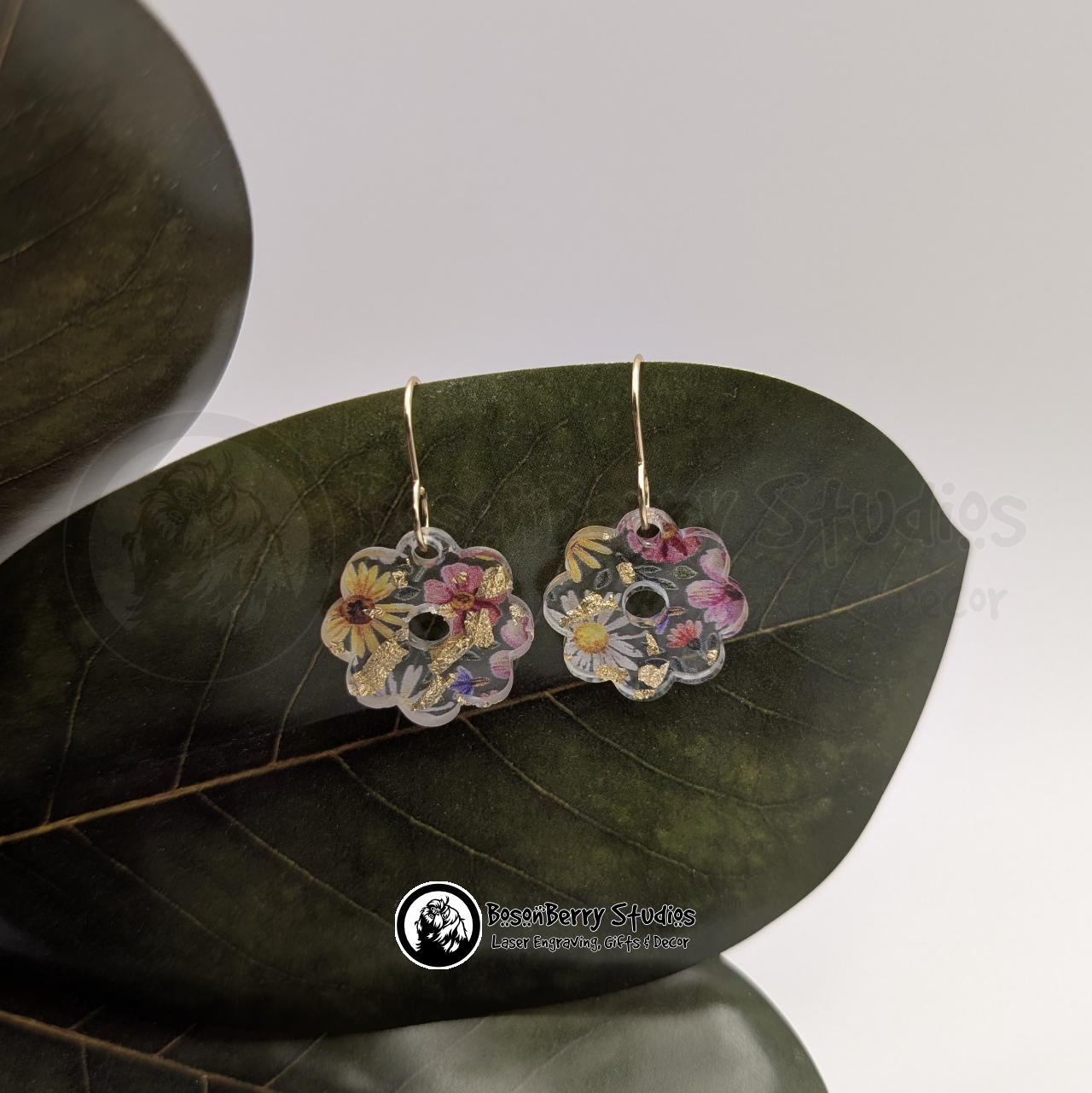 Clear acrylic earrings with embedded gold flakes and a multicolor floral print, in a tiny daisy shape, on a green magnolia leaf in front of a white background with 'BosonBerry Studios' branding.