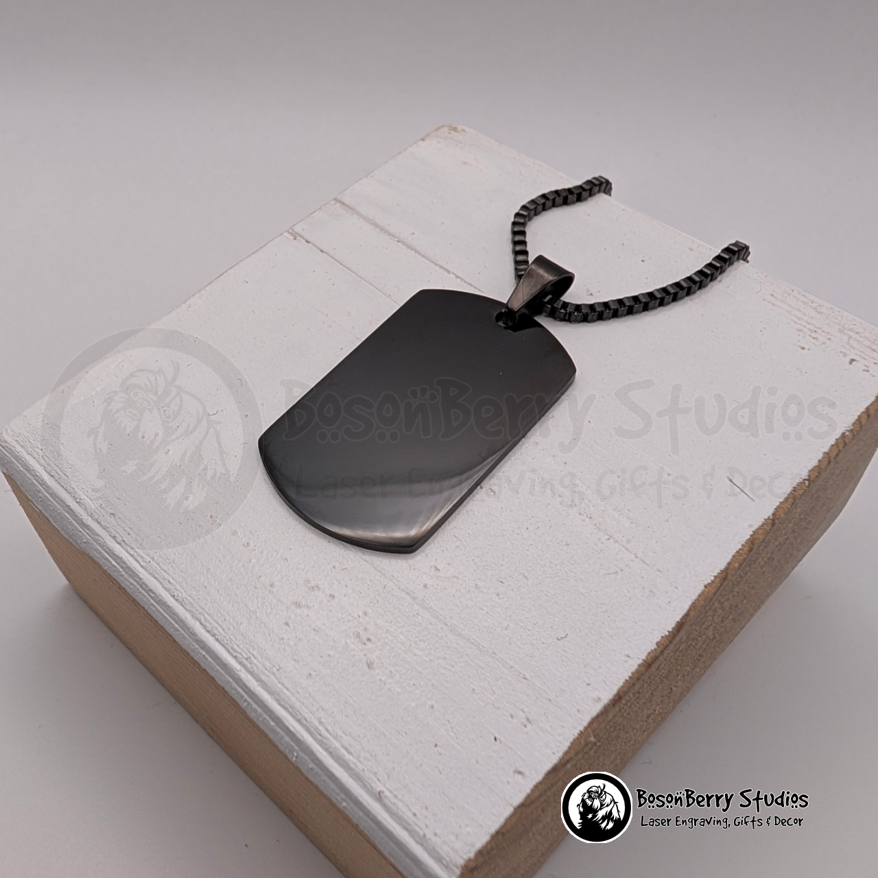 Black dog tag pendant on a white wooden stand with 'BosonBerry Studios' branding.