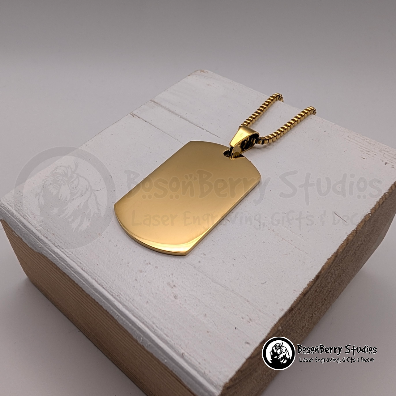 Gold dog tag pendant on a white wooden stand with 'BosonBerry Studios' branding.