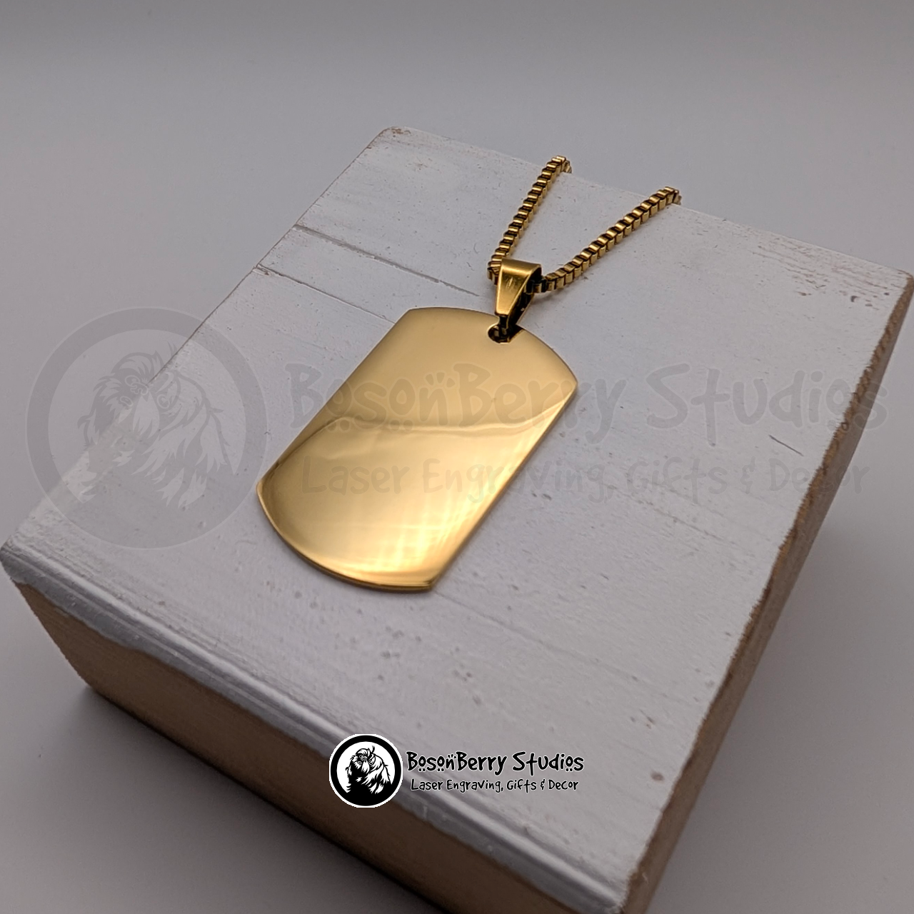 Gold dog tag pendant on a white wooden stand with 'BosonBerry Studios' branding.