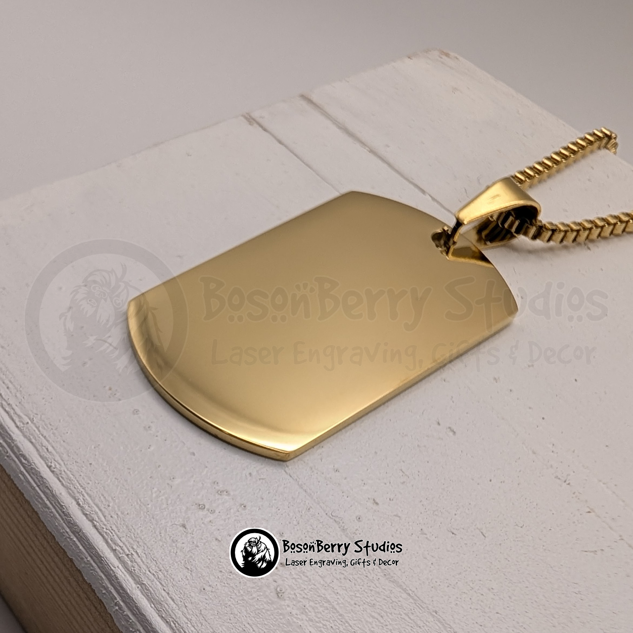 Close up of a gold dog tag pendant on a white wooden stand with 'BosonBerry Studios' branding.