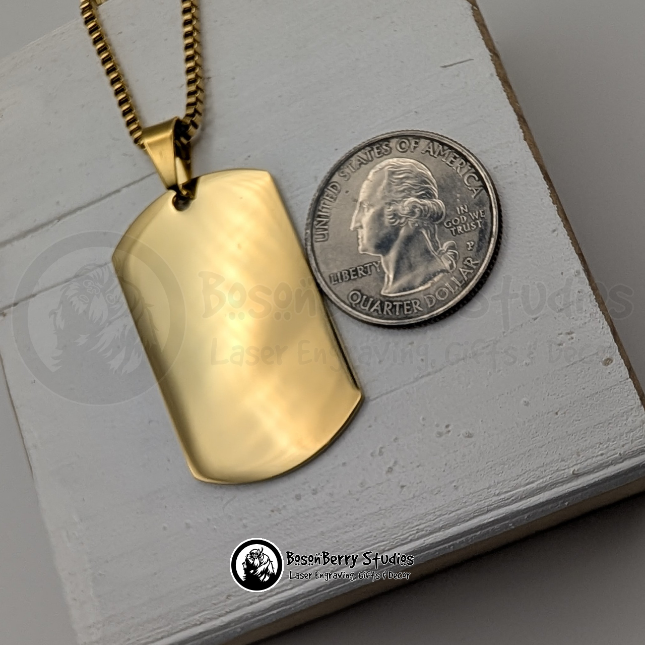 Gold dog tag pendant next to a quarter on a white wooden stand with 'BosonBerry Studios' branding.